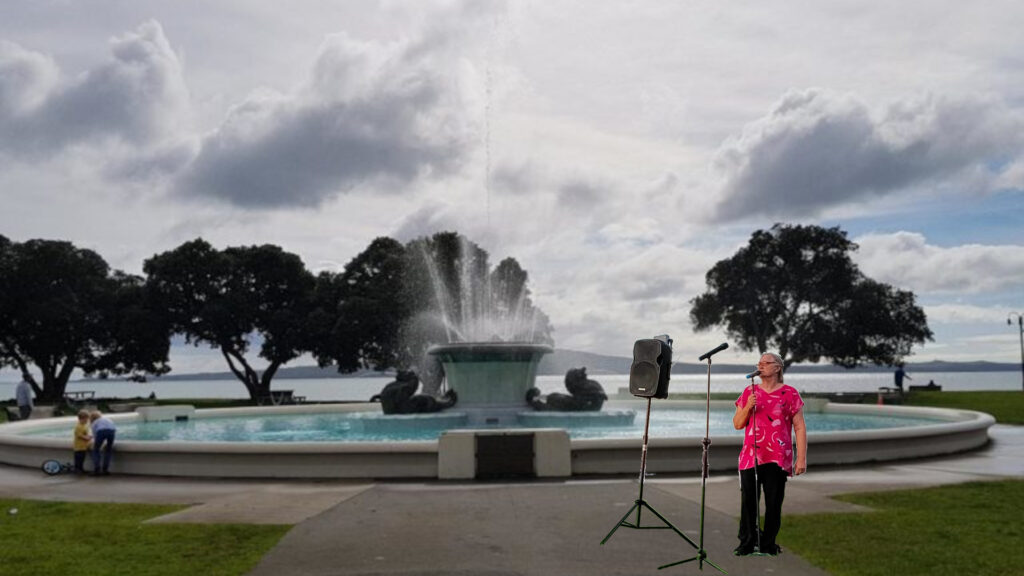 A comedian stands beside a large fountain on the foreshore at Mission Bay.