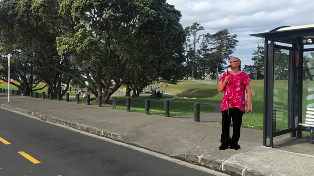 A comedian stands in front of a bus stop outside a park