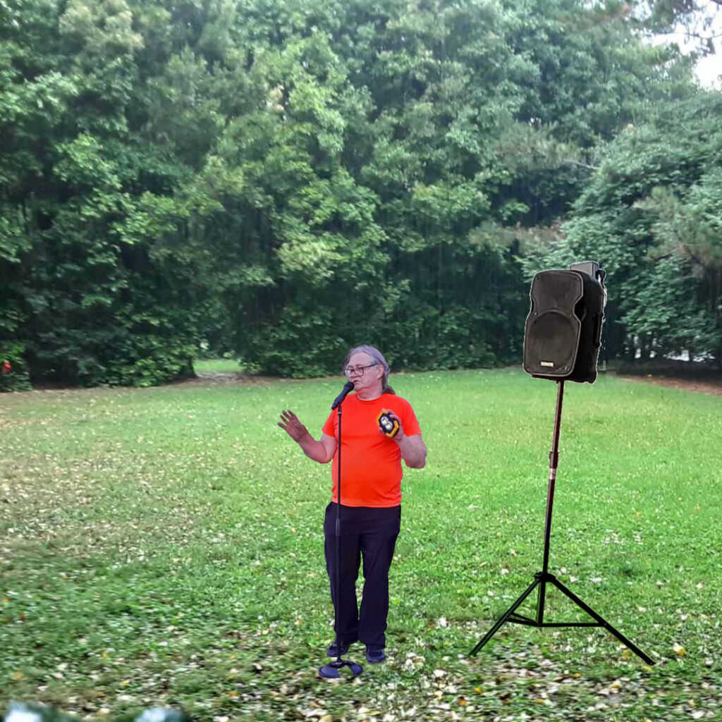 A Comedian stands in a park in front of a loudspeaker.