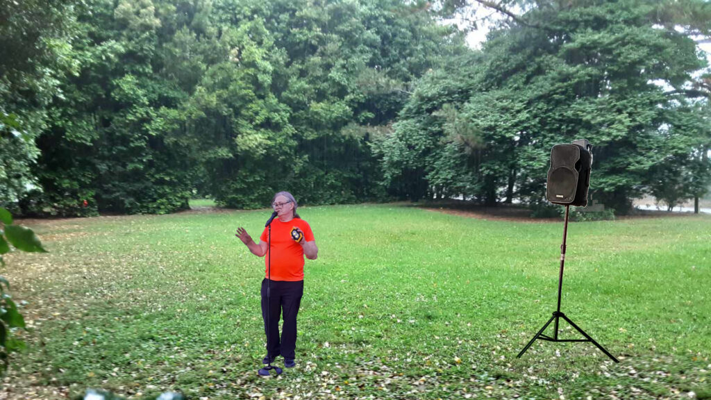 A Comedian stands in a park in front of a loudspeaker.